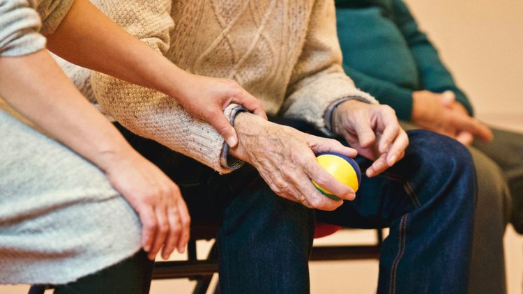 Jüngere Person berührt ältere Person, die einen kleinen Ball in der Hand hält, am Unterarm fest.